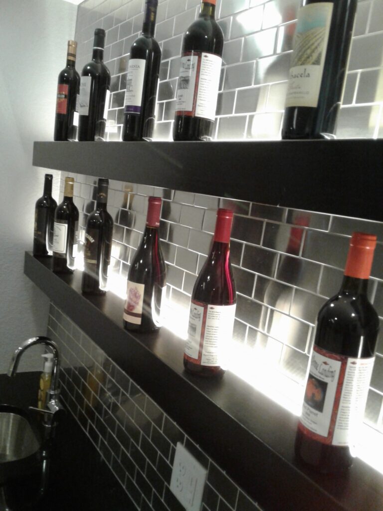 Wine bottles displayed on two wall-mounted shelves with mirrored backsplash, above a black sink with a faucet.