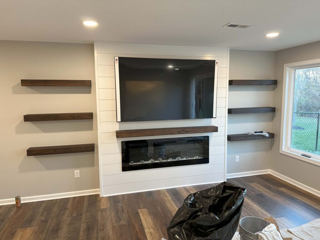 Living room with mounted TV above a modern electric fireplace, surrounded by floating wooden shelves on both sides.