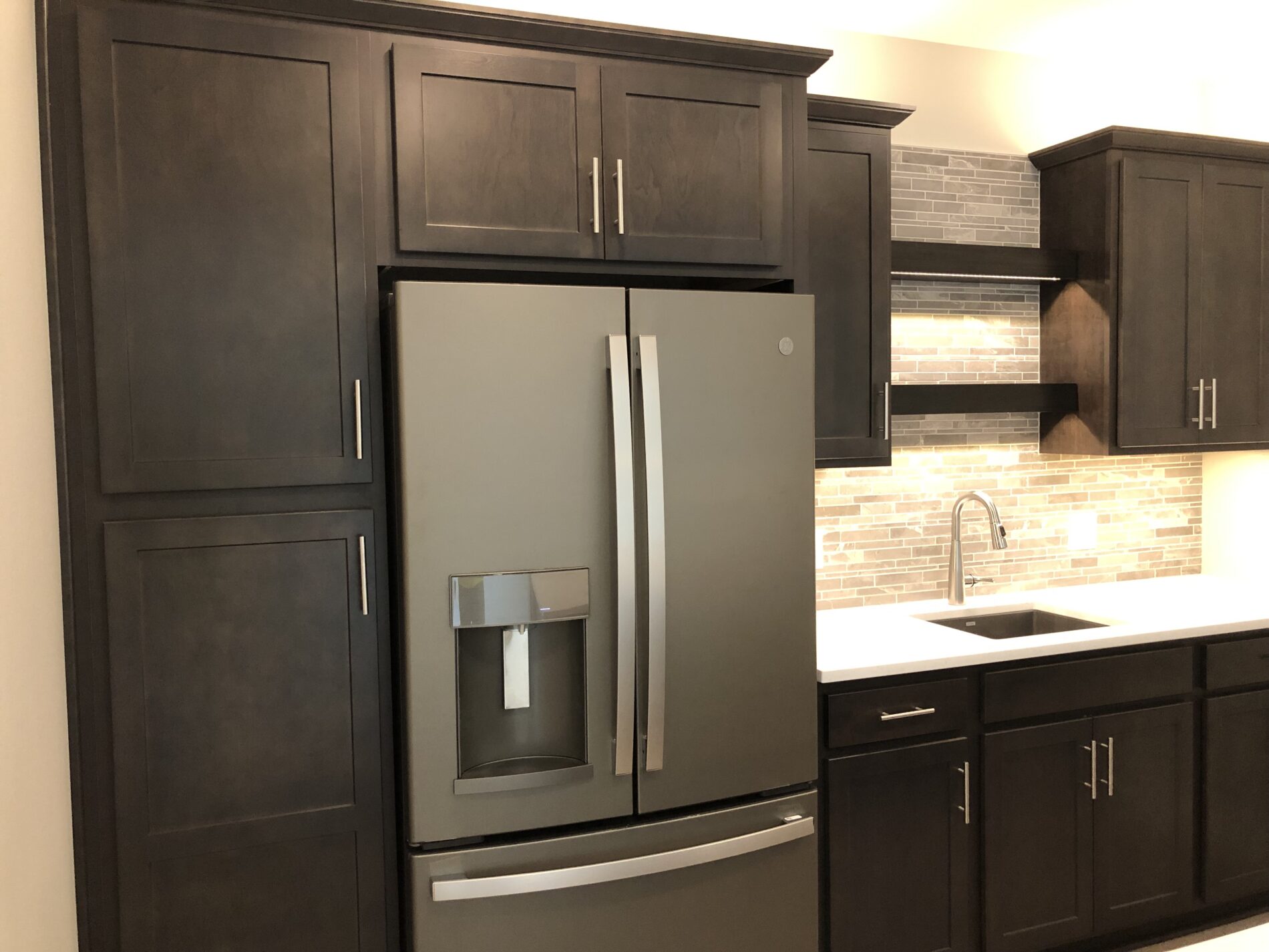 Modern kitchen with dark wood cabinets, a stainless steel refrigerator, and a stone tile backsplash above a white sink.