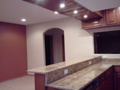 Modern kitchen with granite countertops, wooden cabinets, and recessed lighting, adjacent to an open room with a red accent wall.