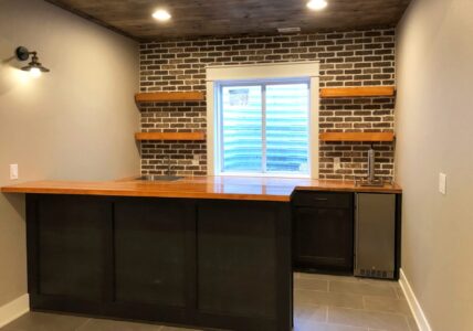 Home bar with a wooden countertop, brick accent wall, open shelves, sink, mini fridge, and a window with a view outside.