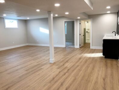 Spacious empty basement with light wood flooring, recessed lighting, a small kitchenette, and an open bathroom entrance.