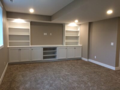 Room with beige carpet, built-in white shelves and cabinets against the wall, overhead recessed lighting.
