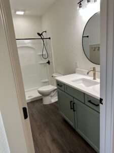 A bathroom with a round mirror, vanity with sink, green cabinets, toilet, and a bathtub with a showerhead.