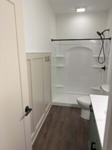 Small bathroom with a shower, toilet, sink, and wood-style flooring. White walls and paneling with a black shower fixture.