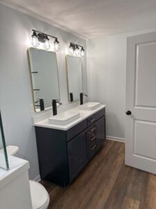 Modern bathroom with dual sinks, black faucets, and mirrors under industrial-style lighting. Wood-look flooring, white walls.