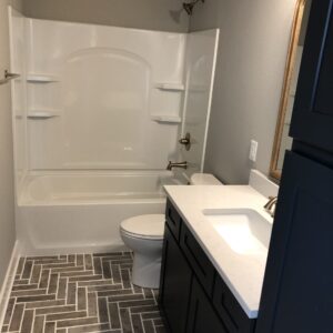 A modern bathroom with a bathtub-shower combo, white sink, dark cabinet, herringbone tile floor, and a large mirror.