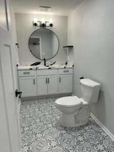 Modern bathroom with round mirror, double sinks, patterned floor tiles, white cabinets, and a toilet.