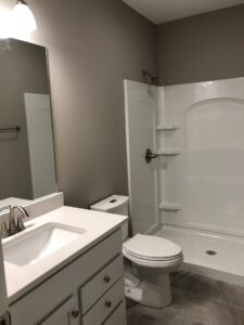 Modern bathroom with a white sink, toilet, and shower area against a gray wall. Simple light fixture above the mirror.