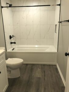 Bathroom with white bathtub and shower, white vanity, and wood-look flooring. Black fixtures and tiled walls.