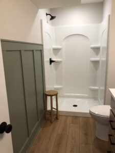 Bathroom with a shower, white walls, green paneling, wood floor, small wooden stool, and a white toilet.