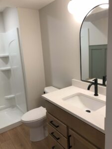A bathroom featuring a shower, toilet, wooden vanity with a white countertop, black faucet, and an arched mirror.