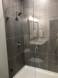 A modern, glass-enclosed shower with gray tile walls and a chrome showerhead, next to a bathroom vanity with a mirror.