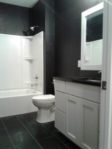 Black and white bathroom with a bathtub, toilet, sink, and mirror. Dark tile flooring and dark walls complement the fixtures.