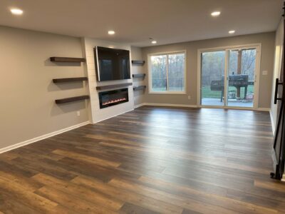 A modern living room with hardwood floors, wall-mounted shelves, a built-in fireplace, and large windows overlooking a patio.