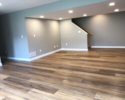 Empty room with wooden flooring, light gray walls, and recessed ceiling lights. A staircase is visible in the corner.